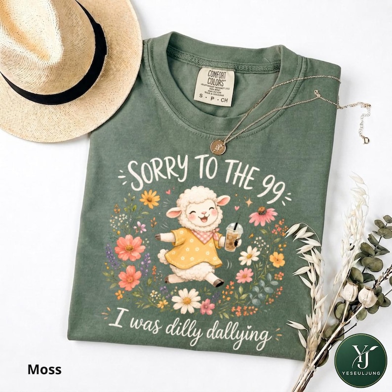 May include: A moss green t-shirt featuring a graphic of a white lamb wearing a yellow polka dot shirt and holding an iced coffee, surrounded by colorful flowers. The text reads SORRY TO THE 99 I was dilly dallying. A straw hat and necklace are present.