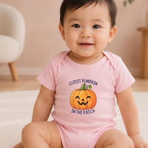 Peut inclure: Un body pour bébé rose clair avec une illustration d'une citrouille orange souriante avec une tige verte. Le texte au-dessus et en dessous de la citrouille indique "CUTEST PUMPKIN IN THE PATCH" en lettres capitales violet foncé.