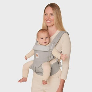 May include: A light gray baby carrier featuring adjustable straps, a structured waist belt, and a padded head support panel. The carrier includes a small logo patch on the front and secure plastic buckles for fastening, designed to hold an infant.