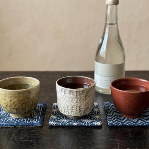 以下が含まれることがあります： 黄色、白、赤の陶器製カップ3つが、白い幾何学模様の青い布製コースターの上に置かれています。白いカップは表面に質感があります。明るいラベルの透明なガラス瓶が、カップの後ろの暗い面に置かれています。
