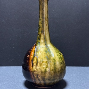 以下が含まれることがあります： 長い細い首と丸みを帯びた底を持つ陶器製の花瓶。釉薬はオリーブグリーン、イエロー、ブラウンの色調が混ざり合ったまだら模様で、光沢のある仕上がりです。表面全体に繊細なひび割れのような質感が施されています。