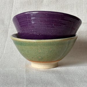 May include: Two stacked ceramic bowls. The top bowl features a deep purple glossy glaze with subtle horizontal texture. The bottom bowl has a light green crackle glaze finish and a visible unglazed beige ceramic base at the bottom.