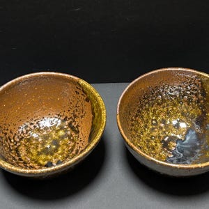 以下が含まれることがあります： 茶色、黄土色、黒の色合いで、質感のあるまだら模様の釉薬が施された2つの丸い陶器のボウル。内面は光沢のある不均一な仕上がりで底に暗い溜まりがあり、外側の縁はざらざらとした土のような質感になっています。
