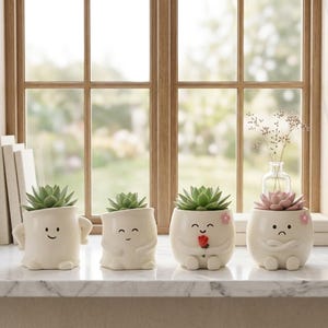 May include: Four off-white ceramic planters shaped like small figures with facial expressions. Each planter holds a green succulent, except for the last one which holds a pink succulent. The figures feature arms, legs, and painted faces.