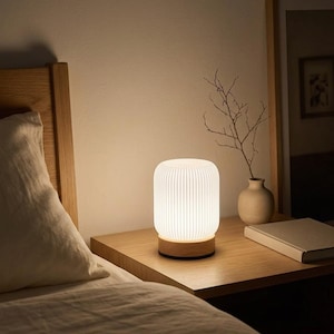 Puede incluir: Una lámpara de mesa cilíndrica con una pantalla de vidrio blanco de rayas verticales y una base de madera clara. La lámpara emite un brillo suave y cálido, iluminando el área circundante con una luz delicada, creando un efecto de calma.