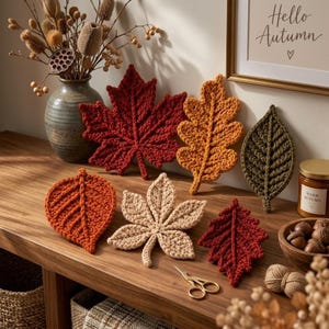 Peut inclure: Une collection de six feuilles au crochet dans diverses couleurs automnales, notamment rouge foncé, orange, beige et vert olive. Les feuilles sont disposées sur une surface en bois avec une petite paire de ciseaux dorés, un vase en céramique et une bougie.