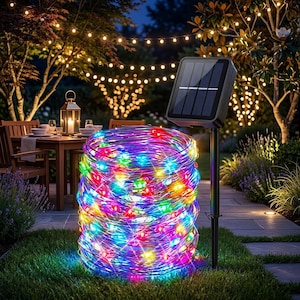 May include: A coiled string of multicoloured LED fairy lights with a black solar panel mounted on a stake. The lights emit a vibrant glow in red, green, blue, and yellow, arranged in a dense cylindrical bundle positioned on a grassy lawn.