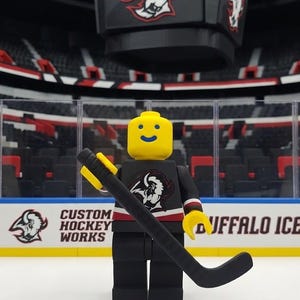May include: A yellow plastic minifigure with a blue smiling face, wearing a black hockey jersey with a white buffalo logo, black pants, and holding a black hockey stick. The figure stands in front of a miniature hockey arena display with the text CUSTOM HOCKEY WORKS and BUFFALO ICE visible on the boards.