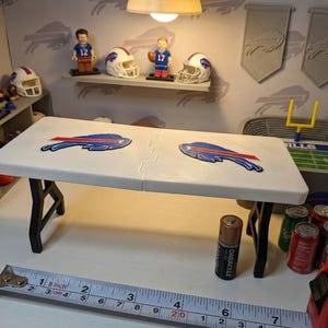 May include: A white miniature folding table with black legs and two Buffalo Bills logos on the surface. A metal measuring tape is positioned in front, showing the table is approximately 7 inches long. The table is set in a sports-themed display.