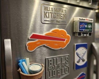 May include: A stainless steel refrigerator door decorated with various Buffalo Bills themed magnets, including a bucket of wings, a KeyBank Center scoreboard, and rectangular plaques reading Bills Mafia's Kitchen Est. 1960 and Bills Mafia's In Big.