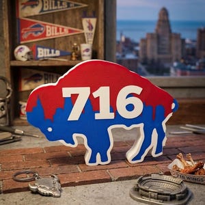 May include: A three-dimensional buffalo-shaped sign featuring a red upper section and a blue lower section with the white number 716 prominently displayed in the center. The sign has a textured surface and white edges, standing upright on a brick surface.