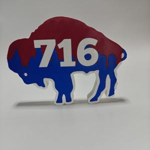 Op de afbeelding: Een wanddecoratie in de vorm van een bizon met een rood bovendeel en een blauw onderdeel, afgewerkt met een witte druppelrand. Het getal 716 staat in een groot, vet wit lettertype in het midden van het getextureerde silhouet van de bizon.
