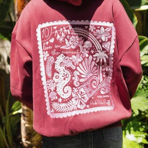 May include: A maroon hoodie featuring a large white rectangular graphic on the back. The design includes a seahorse, tropical flowers, palm trees, and the text "10/ live in the moment" and "with love, Pink Palm Puff" in a scalloped border.