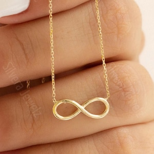 May include: A delicate gold-tone necklace featuring a horizontal infinity symbol pendant suspended from a thin, fine-link chain. The metal has a polished, reflective finish, and the infinity charm is centred on the chain, creating a simple, continuous loop design.
