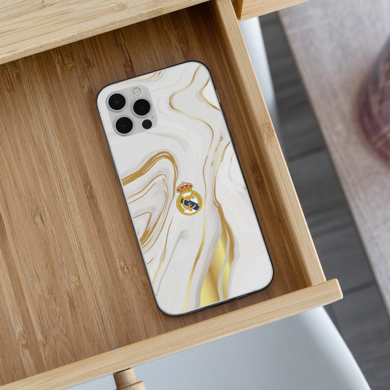 May include: A smartphone case featuring a white and gold marble pattern with swirling veins. Centered on the back is the Real Madrid football club crest, which includes a gold crown, a blue diagonal stripe, and the letters MCF in gold on a white background.