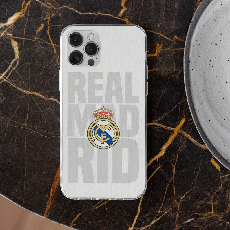 May include: A clear smartphone case featuring a brushed silver background with the text REAL MADRID in large block letters and the official Real Madrid football club crest in the center, displaying a gold crown, blue diagonal stripe, and gold trim.