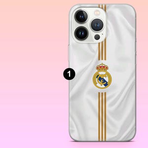 May include: A white smartphone case featuring a silky fabric texture, three vertical gold stripes, and the Real Madrid football club crest centered on the back. The design includes a circular logo with a blue diagonal band and a red crown on top.