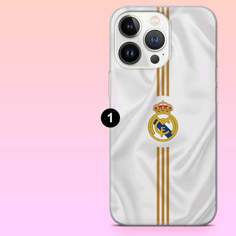 May include: A white smartphone case featuring a silky fabric texture, three vertical gold stripes, and the Real Madrid football club crest centered on the back. The design includes a circular logo with a blue diagonal band and a red crown on top.