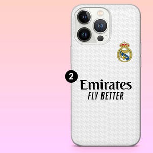 May include: A white smartphone case featuring a subtle light gray houndstooth pattern, the Real Madrid club crest, and the black text Emirates FLY BETTER. A black circle with the number 2 is positioned to the left of the text on the case.
