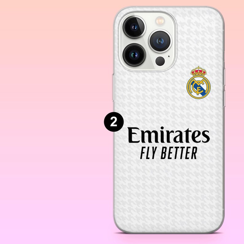 May include: A white smartphone case featuring a subtle light gray houndstooth pattern, the Real Madrid club crest, and the black text Emirates FLY BETTER. A black circle with the number 2 is positioned to the left of the text on the case.