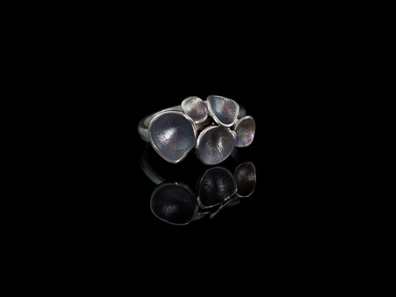 K&ouml;nnte beinhalten: Ein silberner Ring mit einer Gruppe von f&uuml;nf strukturierten, abgerundeten, Bl&uuml;tenblatt-artigen Formen. Der Ring hat eine dunkle, fast schwarze, oxidierte Oberfl&auml;che.