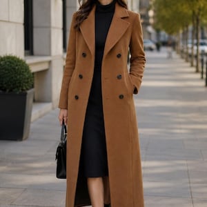 May include: A long, camel-colored double-breasted wool coat with notched lapels and black buttons, worn over a black turtleneck dress and paired with black ankle boots and a black handbag. The coat features a structured fit and side pockets.