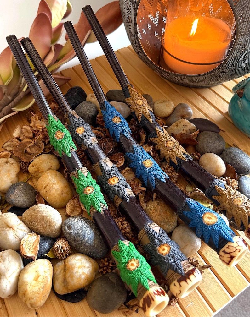 Handcrafted tepi applicator pipes with sculpted leaf collars in green, blue and gold, resting on river stones