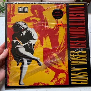 May include: A vinyl record album cover with a yellow and red background. The album cover features a black and white image of a person with a guitar. The album title is "Guns N' Roses Use Your Illusion I" in black text.