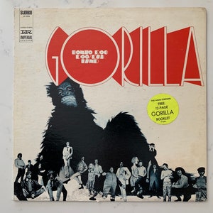 May include: A vintage vinyl record album cover featuring a large black gorilla with a group of people standing in front of it. The album title is "Gorilla" in red letters with the band name "Bonzo Dog Doo-Dah Band" in a smaller circle. The album cover also includes a yellow sticker that says "This album contains free 12-page Gorilla Booklet."