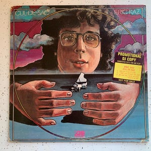 May include: A vinyl record cover featuring a portrait of a man with glasses and a piano in the background. The cover is blue, pink, and yellow with a circle around the man's face. The text on the cover reads "CUL-DE-SAC" and "ERIC KAZ". The text "PROMOTIONAL DJ COPY" is in a white box with black text. The text "SUGGESTED CUTS FOR AIR PLAY" is in a smaller white box with black text. The text "ERIC KAZ CUL-DE-SAC" is in a white box with black text. The text "NOT FOR SALE" is in a white box with black text.