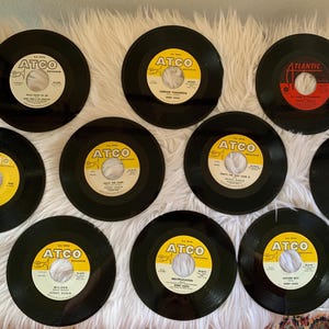 Puede incluir: Diez discos de vinilo negros con etiquetas amarillas. Las etiquetas tienen el texto "ATCO Records" y el título de la canción. Los discos están sobre una superficie blanca y esponjosa.