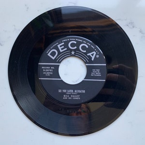 May include: A black vinyl record with a white label featuring the text "DECCA" and the song title "SEE YOU LATER, ALLIGATOR" by Bill Haley and His Comets. The record is a 45 RPM single.