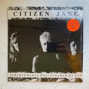 May include: A black and white photo of a record album cover for Citizen Jane. The album cover features three people, two women and one man, with the album title "Citizen Jane" and the song titles "Independence Day" and "Touched By You" printed on the cover.