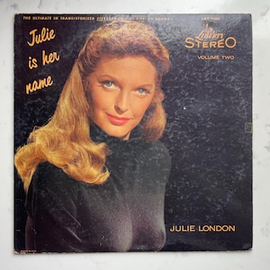May include: Vintage vinyl record album cover titled "Julie is her name" featuring a portrait of Julie London. The album cover has the Liberty Stereo logo and the text "Volume Two". The background is black with a textured appearance.