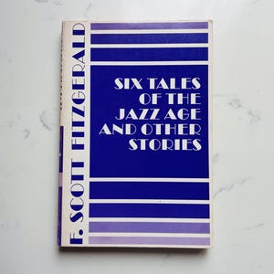 May include: A vintage book titled "Six Tales of the Jazz Age and Other Stories" by F. Scott Fitzgerald. The book cover is a deep blue with white text and horizontal purple stripes. The book is set against a white marble background.