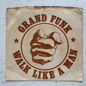 May include: A vintage record label with a brown background and a circle in the center. The circle features a brown hand pointing at the viewer and the text "Grand Funk Walk Like A Man".