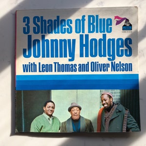 May include: Vintage vinyl record album cover for "3 Shades of Blue" by Johnny Hodges, featuring Leon Thomas and Oliver Nelson. The cover has a blue and white design with a photo of three men. The album is a stereo recording.