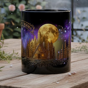 Może przedstawiać: Czarny ceramiczny kubek ze złotą grafiką przedstawiającą chatę w lesie pod wielkim pełnym księżycem. Wzór zawiera szczegółowe korzenie drzew u podstawy, gwiaździste fioletowe nocne niebo oraz napis MOODNEST DESIGNS w rogu kubka.