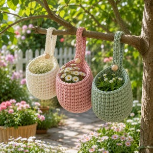 May include: Three hanging crochet baskets in cream, dusty pink, and sage green, each featuring a single wooden bead at the top of the handle. These rounded, textured containers hold small white flowers and are suspended from a tree branch.