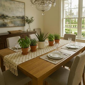 Può includere: Un lungo runner da tavola intrecciato color crema con un motivo a rombi ripetuto e bordi sfrangiati, centrato su un tavolo da pranzo in legno. Il runner è decorato con tre piante verdi in vasi di terracotta e bicchieri di vetro.