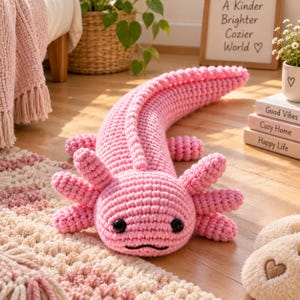 Peut inclure: Une peluche axolotl rose crochetée avec une longue queue incurvée, une tête arrondie et six branchies externes. La créature présente deux yeux en perles noires et une bouche noire cousue. La texture est en laine avec des mailles visibles.