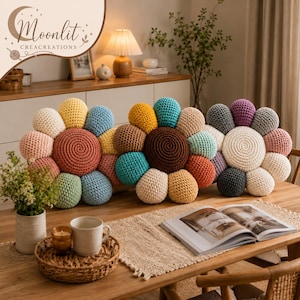 May include: Three flower-shaped crochet pillows featuring multi-colored petals and a central spiral design. The pillows are arranged on a wooden table, showcasing various shades including yellow, blue, pink, brown, and white yarn textures.