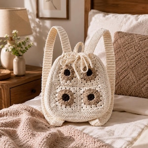May include: A cream-coloured crochet backpack featuring a drawstring closure and four square motifs on the front. Each square motif has a dark brown centre surrounded by light brown and cream yarn. The bag includes two shoulder straps.