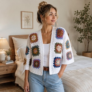 May include: A white crochet cardigan featuring a granny square pattern with multi-colored floral motifs in shades of blue, yellow, pink, and green. The garment has three-quarter length sleeves, an open front, and a ribbed hemline.