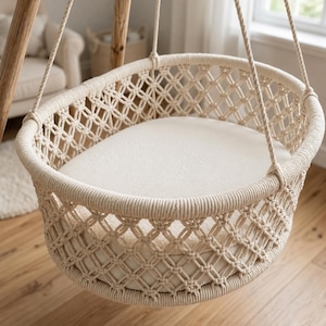 Puede incluir: Una cuna colgante de macramé en color blanco roto con un patrón de diamantes tejidos y un marco de cuerda circular grueso. El interior contiene un colchón liso de color sólido. La cuna está suspendida por cuatro cuerdas gruesas a un marco de madera.