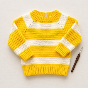 May include: A long-sleeved crochet sweater featuring horizontal stripes in bright yellow and white. The garment has a ribbed crew neckline, cuffs, and hem, with raglan-style sleeves. A dark brown wooden crochet hook is positioned to the right.