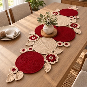 May include: A decorative table runner set featuring crocheted circular mats and floral motifs in deep red and cream yarn. The arrangement includes solid red and cream circles, leaf-shaped accents, and flower-shaped pieces, centered by a white vase.