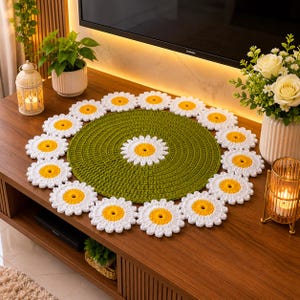 May include: A round, olive green crocheted mat featuring a central white and yellow daisy motif, surrounded by a border of sixteen individual white and yellow crocheted daisy flowers arranged in a circular pattern on a flat surface.