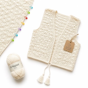 Puede incluir: Un chaleco de ganchillo color blanco roto con un patrón texturizado, que presenta dos borlas largas en la parte inferior y una etiqueta de papel marrón con la inscripción Let's All Crochet. Junto a él hay un ovillo de lana Signet Aran y una pieza tejida.