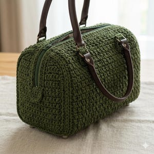 Könnte beinhalten: Eine dunkel olivgrüne Häkeltasche mit einer strukturierten, dicken Garnoberfläche. Die Tasche verfügt über zwei dunkelbraune Kunstledergriffe, die mit Metallringen und Nieten befestigt sind, einen Reißverschluss oben und kleine Metallfüße am Boden.